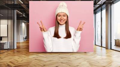 Photo of beautiful brunette woman shows victory or peace gesture, smiles pleasantly, being in high spirit, wears white hat with pompon and sweater, isolated over pink background. Body language Wall mural