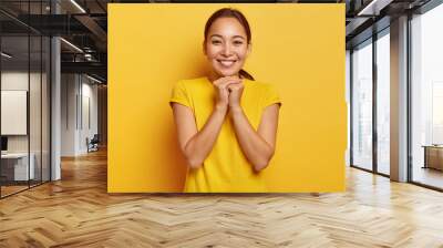 Photo of charismatic Asian female keeps hands together near chin, smiles gently, has cute expression, dark hair combed in pony tail, wears vivid yellow t shirt, entertained in awesome company Wall mural