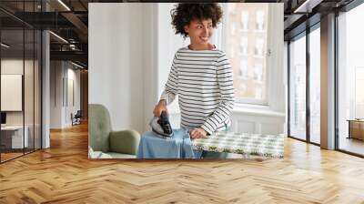 Photo of cheerful black housewife does housework at home during weekend, irons clothes on ironing board, looks joyfully aside. Lovely dark skinned wife irons husbands shirt who is going on work Wall mural