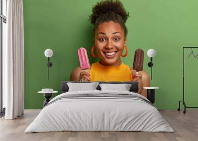 Photo of cheerful dark skinned African American woman holds strawberry and chocolate ice cream smiles broadly dressed in casual yellow t shirt poses against green background. People summer concept Wall mural