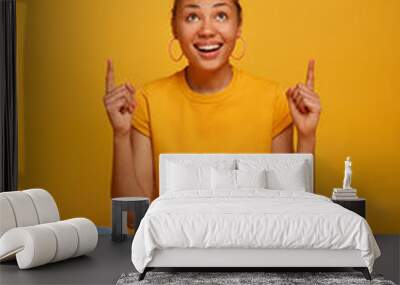 Photo of cheerful ethnic young woman sits on floor crossed legs, points above, shows advertisement, smiles friendly, wears casual yellow t shirt and jeans, participates in interesting discussion Wall mural