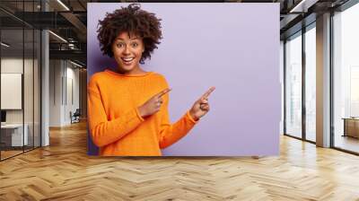 Photo of delighted African American woman points away with both index fingers, promots awesome place for your advertising content, isolated over purple background, gives advice, shows copy space Wall mural