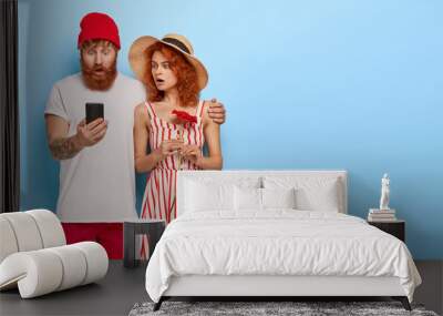 Photo of emotive red haired wife and husband stare at modern smartphone device, react on unexpected comments under their photo in social network, embrace and stand together over blue background Wall mural