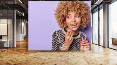 Photo of good looking young woman with curly hair applies lip gloss wears bright makeup undergoes beauty procedures before dating isolated over purple background blank space for your promotion Wall mural