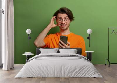 Photo of handsome hipster guy with trendy hairstyle, scratches head and thinks about something, uses modern mobile phone, checks newsfeed, dressed in orange t shirt, isolated on green background Wall mural