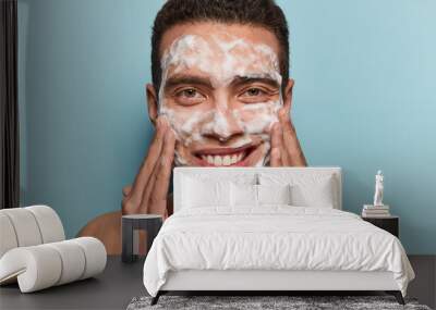 Photo of handsome satisfied European man cleans and washes face with soap, enjoys purity of skin, smiles gently, has pampering procedure, satisfied with skin condition. Rejuvenation concept. Wall mural