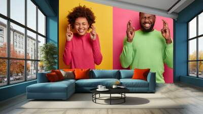 Photo of hopeful lucky Afro America woman and man cross fingers for good luck, believe fortune will come, hope wish fullfill, anticipate miracle happened, pose against yellow and pink background Wall mural