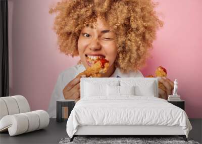 Photo of hungry young European woman eats delicious fried chicken nuggets with ketchup winks eye enjoys eating fatty appetizing unhealthy food dressed in formal clothes isolated over pink background Wall mural