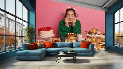 Photo of lovely millennial girl holds knife and fork, thinks what to eat first, sits at big table full of cookies, pastries and cakes, eats different desserts during holidays, has bad eating habits Wall mural