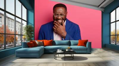 Photo of overjoyed Afro American man laughs at funny story, cant stop giggling, has white teeth, thick beard, wears formal suit, squints face, isolated on pink background, glad to achieve success Wall mural