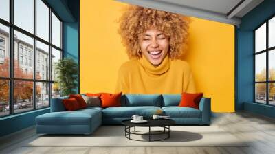 Photo of overjoyed European woman laughs sinecerely smiles broadly has upbeat mood keeps eyes closed wears casual warm jumper isolated over yellow background. Authentic happy emotions concept Wall mural