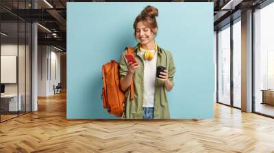 Photo of positive young woman in casual clothes, downloads multimedia file on cell phone, has headphones on neck, holds disposable cup with cappuccino, carries backpack, isolated on blue wall Wall mural