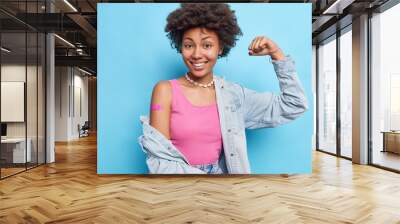 Photo of satisfied curly haired woman raises arm shows biceps wears pink t shirt denim shirt necklace adhesive bandage on shoulder after vaccination feels good and protected encourages to vaccinate Wall mural