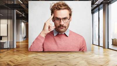 Photo of serious unshaven young male wears round spectacles, keeps finger on temple and looks strictly, tries to concentrate and make important speech, isolated over white concrete background Wall mural