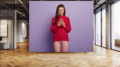 Photo of smiling Caucasian teenage girl focused in modern cell phone, enjoys online communication, wears long knitted red sweater, sends text messages to friend, stands over purple background Wall mural