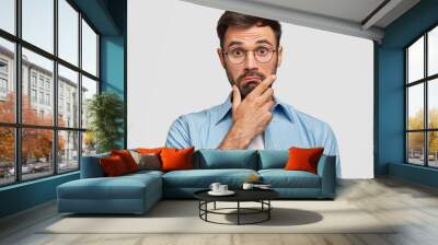 Photo of surprised bearded male holds chin and looks in hesitation, wonders latest news, wears spectacles and elegant blue shirt, isolated over white background. People and facial expressions concept Wall mural