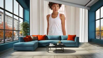 Photo of thoughtful dark skinned girl wears white casual t shirt, stands near window indoor during rainy day, has pensive look aside, enjoys leisure time. Horizontal view of African American lady Wall mural