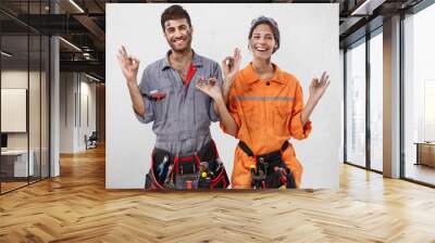 Picture of young female and male workers of maintenance service wearing working clothes and waist bags with tools, smiling, making mudra gesture, trying to stay clam and positive about their hard work Wall mural