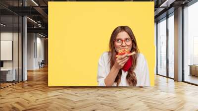 Pleasant looking female enjoys delicious slice of pizza, being hungry, looks with dreamy expression upwards, likes this taste, wears eyewear and white oversized shirt, stands against yellow background Wall mural