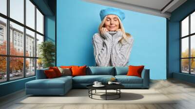 Pleased adult woman wears comfortable knitted sweater keeps hands on collar stands with closed eyes enjoys peaceful moment isolated over blue background. Satisfied fashionable middle aged lady Wall mural