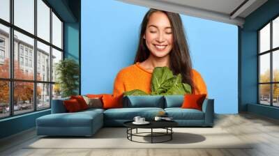 Pleased Asian woman with dark long hair holds green bok choy vegetable, going to make tasty eco salad, keeps eyes shut, wears orange jumper, has healthy eating, isolated over blue background Wall mural