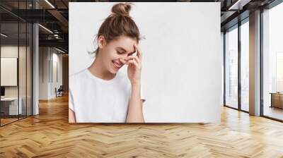 Pleased beautiful female model with hair knot looks with admiration, pleasant smile down, has shy expression as recieves compliment from boyfriend, reveals real good feelings, isolated on white Wall mural