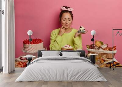 Pleased brunette woman holds small creamy muffin, bakes many desserts for New Year holiday or festive time, demonstrates her culinary abilities, has special occasion, degustates homemade cupcake Wall mural