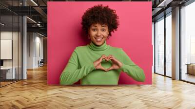 Pleased cheerful dark skinned Afro American woman shapes heart, confesses in love and expresses sympathy, smiles broadly, shows white teeth, wears green turtleneck, isolated on pink background Wall mural