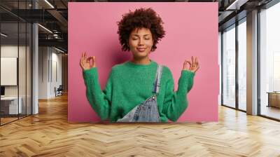 Pleased curly haired woman tries to calm down, unites with nature, raises hands and shows zen gesture, meditates or does yoga indoor, closes eyes, enjoys peaceful atmosphere for good relaxation Wall mural