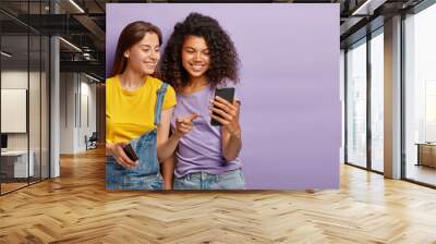 Pleased female friends watch something interesting on mobile phone, have happy relaxed expressions, focused in device, enjoy using modern technologies, isolated over purple wall with empty space Wall mural