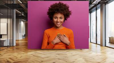 Pleased girl in orange jumper presses palms to heart, makes thankful gesture, touched with cordial congratulations, smiles positively, isolated over purple background. Acknowledgement concept Wall mural