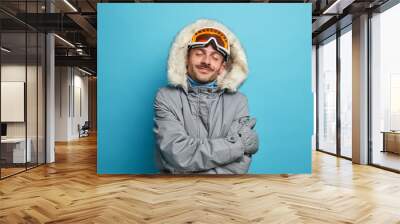 Pleased male snowboarder feels comfort and warm in winter jacket embraces himself recalls nice moment of going skiing during nice cold day stands with closed eyes isolated on blue background Wall mural