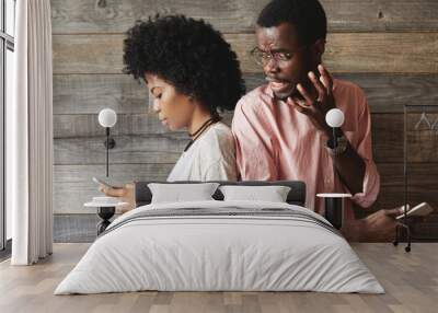 Portait of annoyed African man in glasses angry with his attractive girlfriend who is absorbed in surfing Internet and texting friends via social networks while standing back-to-back with her Wall mural