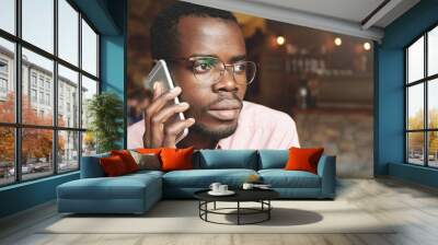 Portrait of African corporate worker wearing pink shirt talking on mobile phone with serious expression while sitting at a coffee shop against wooden wall background. Lifestyle and people concept Wall mural