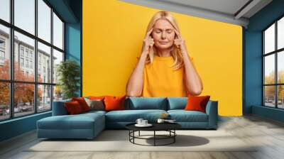 Portrait of attractive senior lady stands with eyes closed keeps fingers on temples thinks about something and tries to find solution has bad memory dressed in casual wear isolated on yellow wall Wall mural