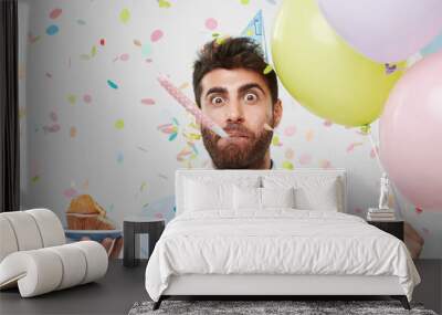 portrait of bearded man wearing party hat and shirt, whistling in party horn, holding cupcake in han Wall mural