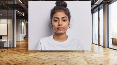 Portrait of beautiful serious brunette woman focused at camera has dark hair combed in bun dressed in casual t shirt isolated over white background. Confident Idian female model with calm expression Wall mural