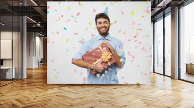 Portrait of cheerful joyful young Caucasian man with beard enjoying his birthday party, standing isolated in light room with confetti falling down on him, smiling, holding present in beautiful paper Wall mural