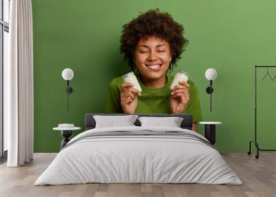 Portrait of cheerful woman holds two bottles of natural dairy yogurt for breakfast, bites lips and has temptation to eat, stands with closed eyes, isolated on green wall. Healthy nutrition concept Wall mural