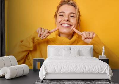 Portrait of cheerful young woman smiles broadly indicates index fingers at face wears casual jumper expresses positive emotions isolated over vivid yellow background. Look at my brilliant smile Wall mural