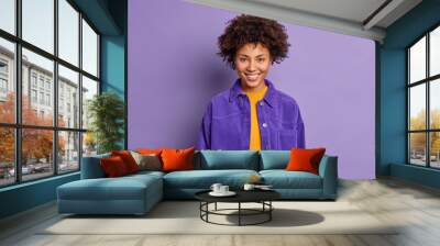 Portrait of curly haired beautiful woman smiles happily wears fashionable purple jacket in one tone with background feels delighted poses glad indoor talks to coworker. Positive emotions concept Wall mural