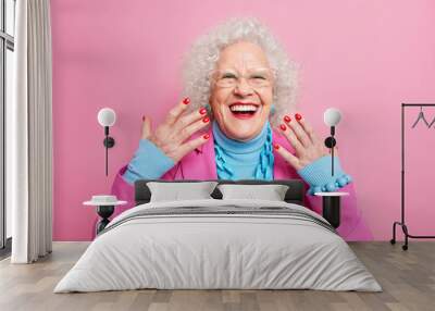 Portrait of elderly curly haired woman raises hands smiles broadly wears spectacles fashionable outfit necklace being in good mood isolated over pink background. Old people and style concept Wall mural