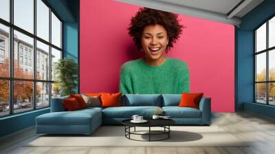 Portrait of good looking curly ethnic woman smiles broadly, enjoys day off, has happy talk with interlocutor, discusses holiday preparation, wears green sweater, isolated on pink background. Wall mural