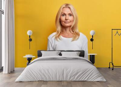 Portrait of good looking middle aged woman with wrinkled face natural beauty blonde hair looks directly at camera has calm expression dressed in white casual jumper isolated on yellow studio wall Wall mural