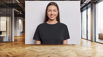 Portrait of good looking young woman with no makeup natural beauty has long dark straight hair dressed in casual black t shirt poses against white background poses in studio for making photo Wall mural