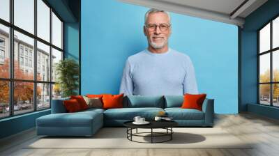 Portrait of handsome bearded European man with grey hair and beard smiles pleasantly looks directly at camera being in good mood has lucky day wears spectacles and sweater isolated over blue wall Wall mural