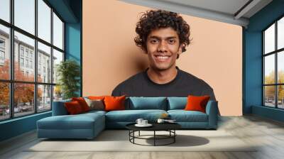 Portrait of handsome cheerful man with curly hair smiles toothily poses happy against brown background dressed casually isolated over brown background. Positive human emotions and ethnicity concept Wall mural