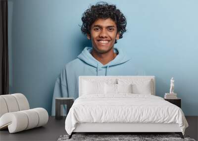 Portrait of handsome curly haired young Hindu man smiles pleasantly dressed in casual hoodie expresses positive emotions poses against blue studio background. Monochrome shot. Youth concept. Wall mural