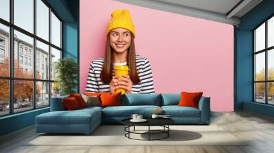 Portrait of happy brunette woman holds takeaway coffee, wears yellow hat and casual striped jumper, looks aside with smille, isolated over pink background. Pretty girl with beverage, has spare time Wall mural