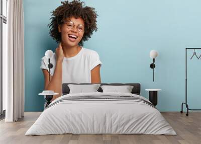 Portrait of happy carefree woman touches neck gently, demonstrates natural beauty, dressed in casual t shirt, smiles broadly, stands over blue background with empty space for your advertisement Wall mural